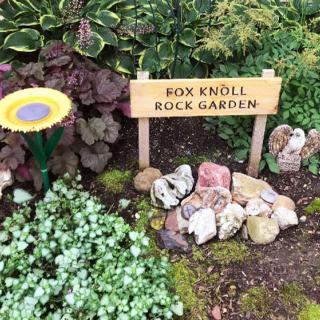 A small rock garden with a sign reading 'Fox Knoll Rock Garden', surrounded by various plants.