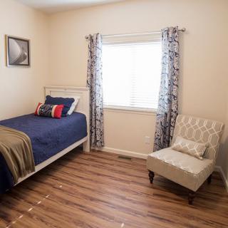 A cozy bedroom featuring a single bed with blue bedding, a comfortable chair, and a window with patterned curtains.