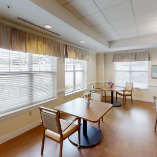 A bright dining room with tables and chairs, featuring large windows and curtains.