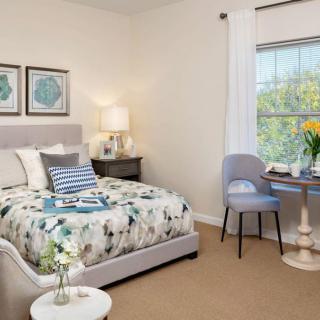 A cozy bedroom with a neatly made bed, bedside lamp, small round table with a chair by the window, and decorative elements creating a tranquil atmosphere.