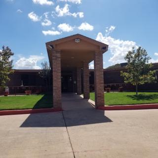 A welcoming entrance of a school building surrounded by green grass and trees under a blue sky.