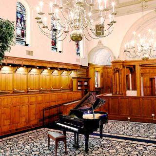A grand piano sits elegantly in a tastefully decorated room featuring chandeliers, wood paneling, and stained glass windows.