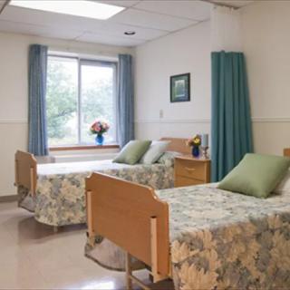 A hospital room with two beds, decorated with floral bedspreads and green pillows, featuring a window with blue curtains and vases of flowers.