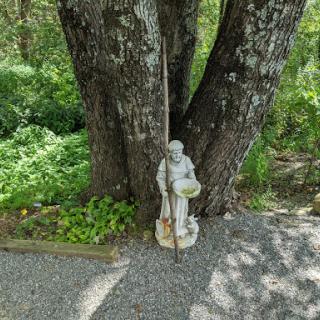 A serene stone statue stands beside a large tree, surrounded by lush greenery.
