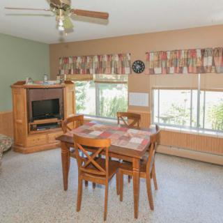 A cozy dining room with a wooden table and chairs, carpeted floor, and large windows adorned with curtains, featuring a ceiling fan and a fireplace in the corner.