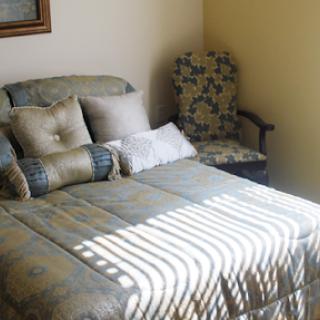 A cozy bedroom featuring a neatly made bed with decorative cushions and a floral armchair, illuminated by sunlight.