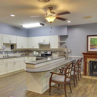 A modern kitchen interior with white cabinets, a countertop with bar stools, a ceiling fan, and wood flooring next to a fireplace.