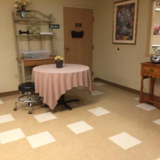 A quiet waiting room with a round table covered by a pink tablecloth, a chair, decorative plants, and artwork on the walls.