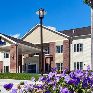 A modern two-story building with a covered entrance, surrounded by purple flowers under a clear blue sky.