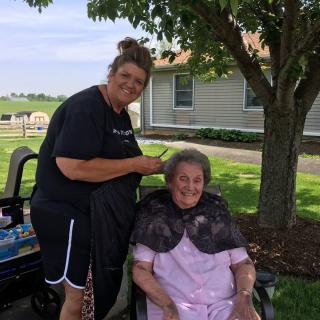 A woman giving a haircut to an elderly person under a tree outdoors.