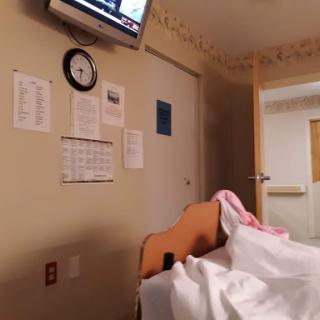 A view of a hospital patient room, featuring a bed, a television displaying news, and a wall clock.