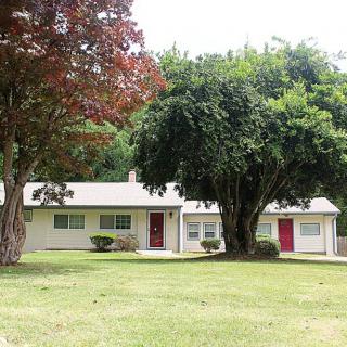 A small white house with red doors surrounded by large trees and a green lawn.