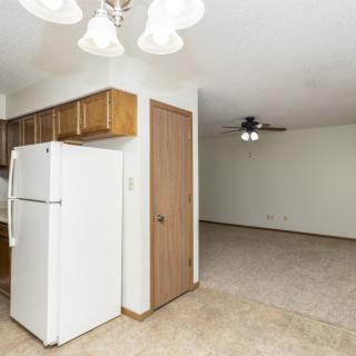 A view of a kitchen with wooden cabinets and a white refrigerator, leading into an open living room with a ceiling fan.