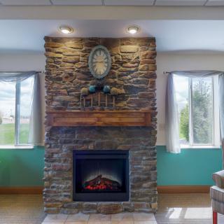 A cozy stone fireplace with a wooden mantle, flanked by two windows in a well-lit room.