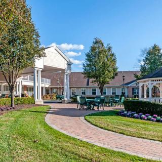 A serene outdoor view of a well-maintained garden with a pathway, trees, a gazebo, and a building in the background.