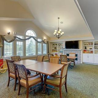 A spacious dining area with large windows, elegant tables and chairs, a chandelier overhead, and a cozy bookshelf with a TV.