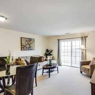 A well-furnished living room with a sofa, chairs, and a dining table, complemented by artwork on the wall and large windows allowing natural light.