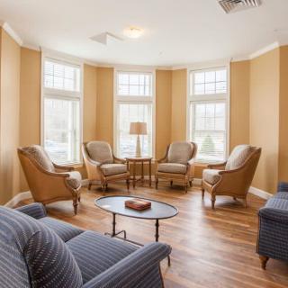 A cozy living room with armchairs arranged around a central table, illuminated by natural light from large windows.