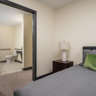 A cozy bedroom with a gray bedspread, a nightstand with a lamp, and an adjoining bathroom.