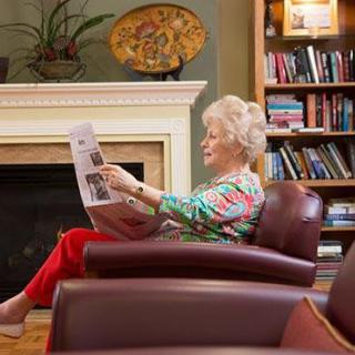 An elderly woman reads a newspaper in a cozy living room setting.