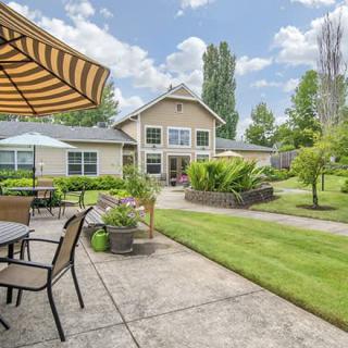 A beautiful backyard with a well-maintained lawn, patio furniture under a striped umbrella, and lush greenery surrounding a modern house.