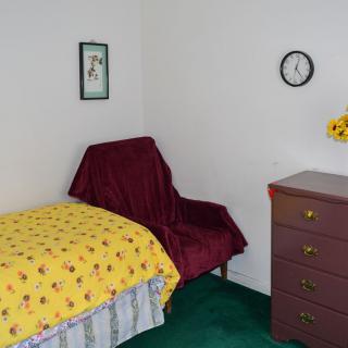 A simple bedroom with a single bed, a small dresser, and a chair, featuring minimal decor including a wall clock.