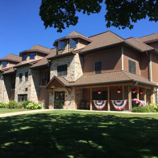 A large residential building with a stone facade and American flags adorning the porch, surrounded by a lush green lawn and trees under a clear blue sky.