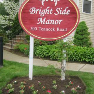 The sign for Bright Side Manor, located at 300 Teaneck Road, surrounded by a well-maintained garden.
