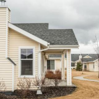 A suburban residential house with beige siding and a small front porch, set against a cloudy sky.