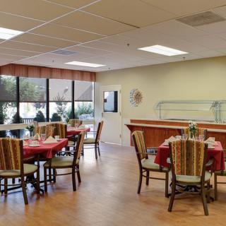 A well-lit dining room with neatly arranged tables and chairs, featuring large windows and a buffet counter.