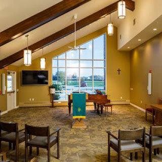 A modern chapel interior with wooden beams, seating, a piano, and a large window providing an outdoor view.