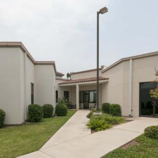 The image shows the exterior of a modern building with a paved pathway leading to the entrance, surrounded by well-maintained landscaping.