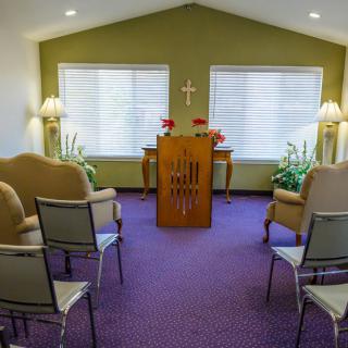 A small chapel interior with seating, plants, and an altar under a cross.