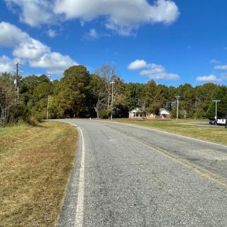 A quiet rural road winding through a countryside landscape under a bright blue sky with scattered clouds.