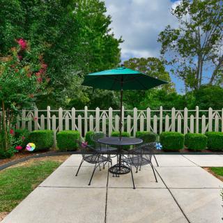 A peaceful patio area with a table and chairs under a green umbrella, surrounded by a garden and a picket fence.