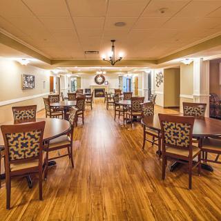 A spacious dining room with wooden flooring, elegantly arranged tables and chairs, illuminated by a centered chandelier.