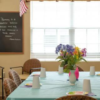 A dining room with a table set for a gathering, featuring a bouquet of flowers and a chalkboard in the background.