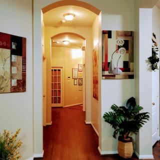 A warmly lit hallway adorned with paintings and plants, featuring a wooden floor leading to a door.