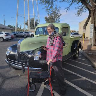 An elderly person stands next to a vintage green truck in a parking lot.