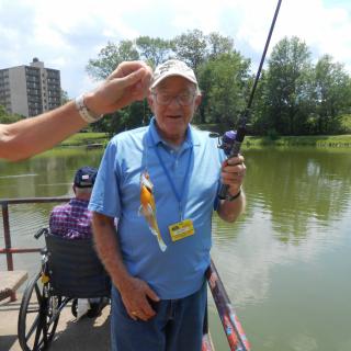 An elderly man is fishing by a lake on a sunny day, holding a fishing rod and wearing a hat. Another person in a wheelchair can be seen nearby.