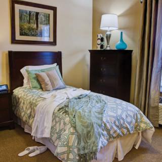 A cozy bedroom featuring a single bed with decorative pillows and a patterned blanket, accompanied by a dresser with a lamp and artwork on the wall.