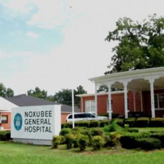 The image shows the exterior of Noxubee General Hospital with a sign in the foreground.