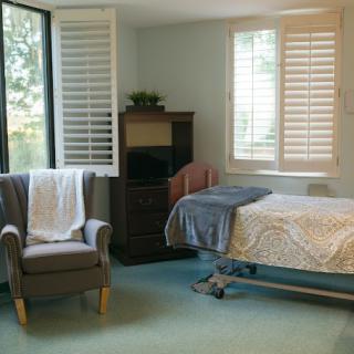 A cozy bedroom with a hospital bed, chair, and large windows featuring shutters.
