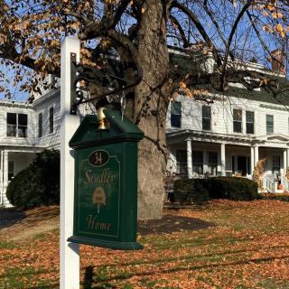 A white house with a green sign in front, surrounded by autumn leaves and trees.
