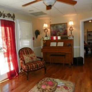 A cozy living room featuring a piano, patterned chair, and warm wooden flooring with red curtains and decorative lamps.