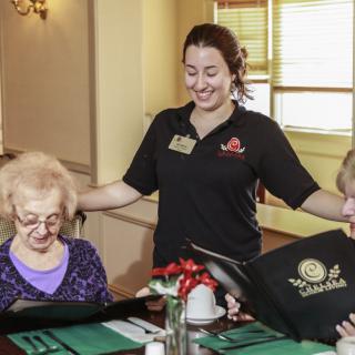 A waitress cheerfully engages with elderly diners as they look at their menus in a restaurant setting.