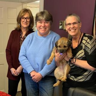 Three women pose together with a small dog in a cozy indoor setting.