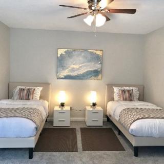 A modern bedroom featuring twin beds with neutral-toned bedding, complemented by a ceiling fan and abstract artwork on the wall.