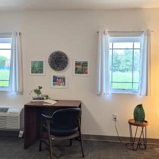 A minimalist workspace featuring a desk between two windows, adorned with small artworks and a table lamp.