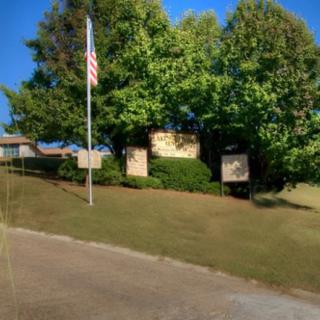 An outdoor scene featuring a sign next to a flagpole with trees and a building in the background.